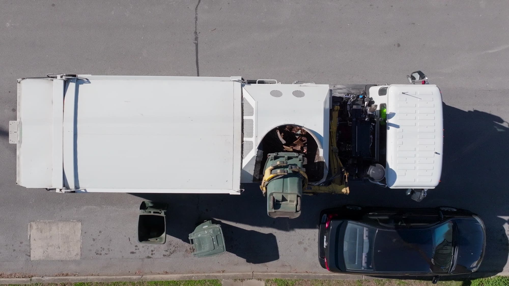 A refuse collection truck seen from directly above, lifting a wheelie bin at the kerb
