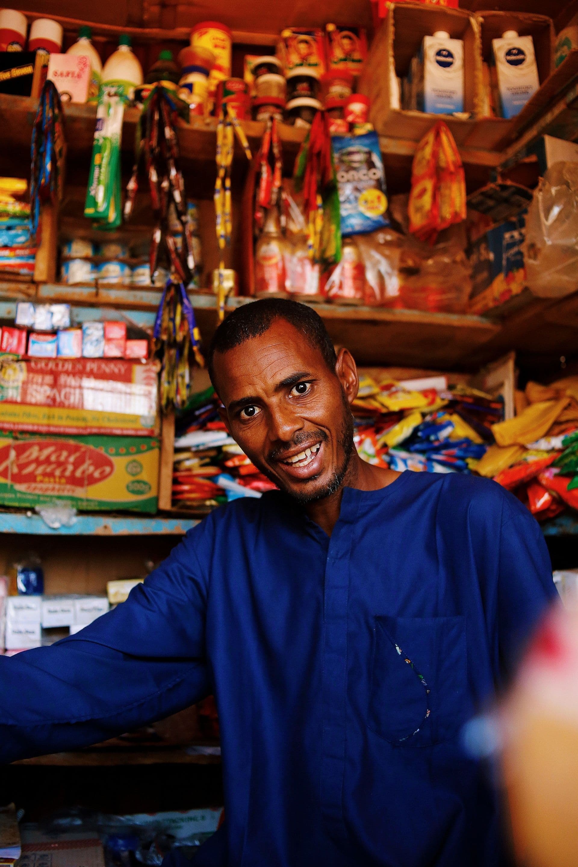 A shopkeeper smiling behind the counter of a small, well-stocked grocery shop